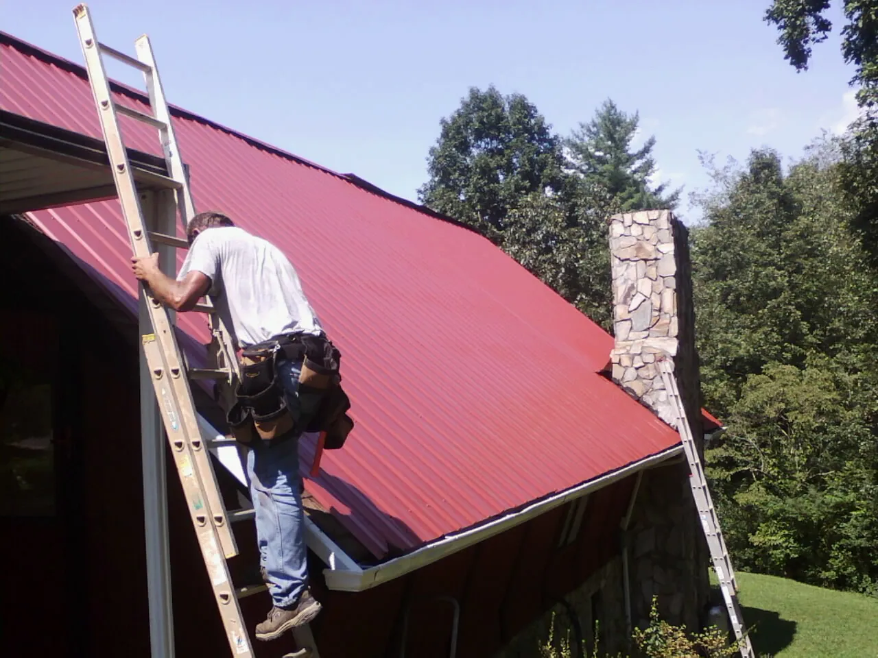 Roofer on a ladder beside a red metal roof and stone chimney — Soffit & Fascia Repair by Roofing Brothers in Pemberville